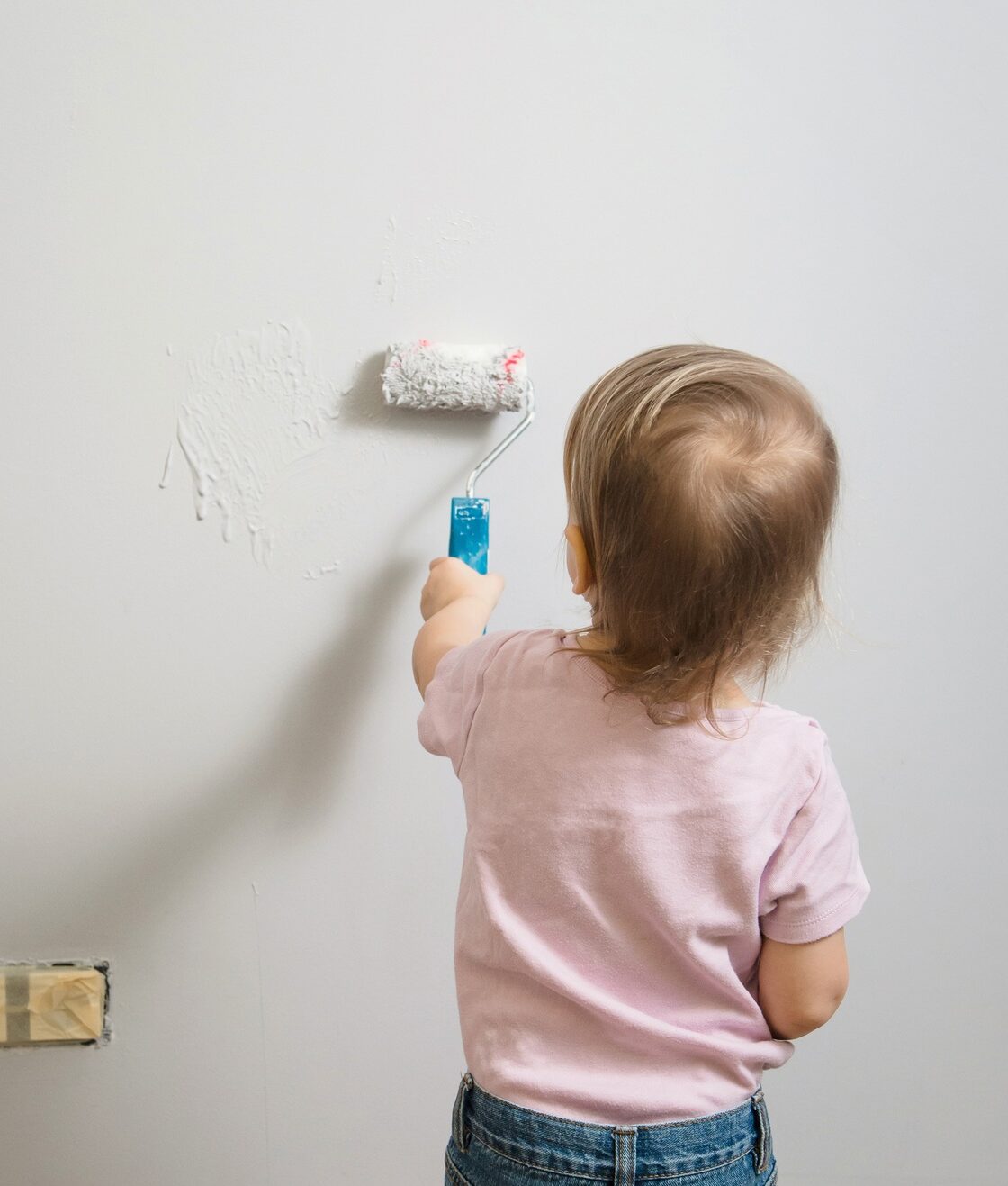 Little child painting the wall with roller in the empty room, grey background. Interior decoration,